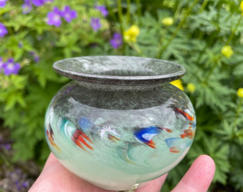 K&ouml;nnte beinhalten: Eine kleine, runde, gr&uuml;ne Glasvase mit einem dunkelgrauen Rand. Die Vase hat ein wirbelndes Muster aus Wei&szlig;, Blau und Orange.