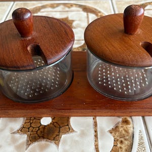 以下が含まれることがあります： 木製の蓋と木製のベースが付いた、ガラス製のスパイスジャーのセット。濃い茶色の蓋には小さなハンドルとスプーン用のスロットがあります。ジャーは長方形の木製トレイに置かれています。