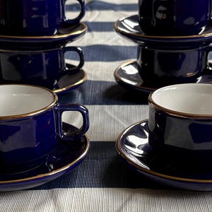 以下が含まれることがあります： 深い青色のセラミック製ティーカップとソーサーの6客セット。カップの内側は白く、カップの縁とソーサーの外縁には細いメタリックゴールドの縁取りが施されています。カップには四角い形状の持ち手が付いています。