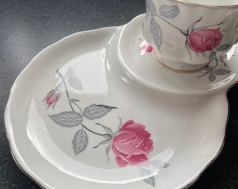 May include: A white teacup and saucer set with a delicate floral design featuring pink roses and gray leaves. The saucer has a unique shape with a raised section for the cup.