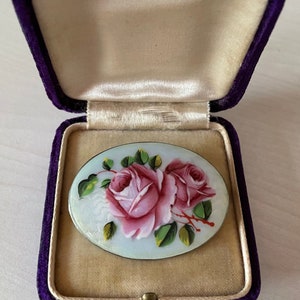 May include: A vintage oval-shaped brooch with a hand-painted design of two pink roses with green leaves on a light blue background. The brooch is in a purple velvet box.