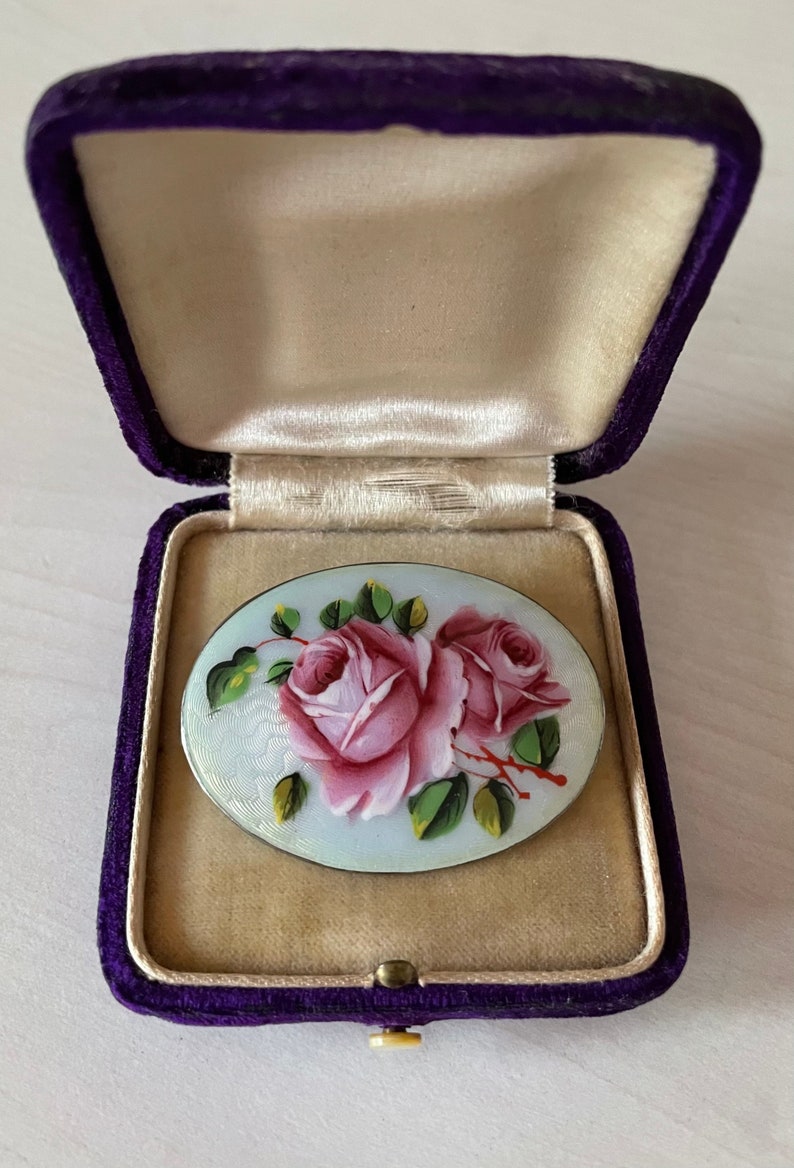 May include: A vintage oval-shaped brooch with a hand-painted design of two pink roses with green leaves on a light blue background. The brooch is in a purple velvet box.