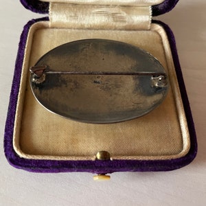 May include: The reverse of a silver oval brooch with a pin and a marking that reads "925". The brooch is in a purple velvet box.
