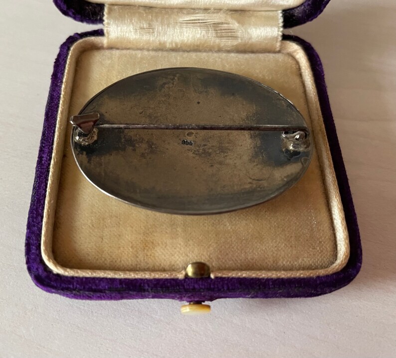 May include: The reverse of a silver oval brooch with a pin and a marking that reads "925". The brooch is in a purple velvet box.