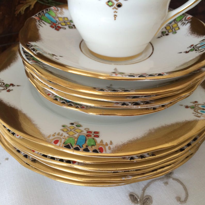 Może przedstawiać: Stos białych porcelanowych talerzy z pozłacanymi krawędziami i kolorowymi geometrycznymi wzorami. Talerze są ułożone w kształcie piramidy, z największym talerzem na dole i najmniejszym talerzem na g&oacute;rze.