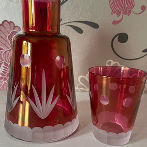 May include: A set of two ruby red glass decanters with etched white designs. The larger decanter has a wide mouth and a tall, slender body. The smaller decanter is a tumbler with a rounded body. Both decanters have a frosted white rim.