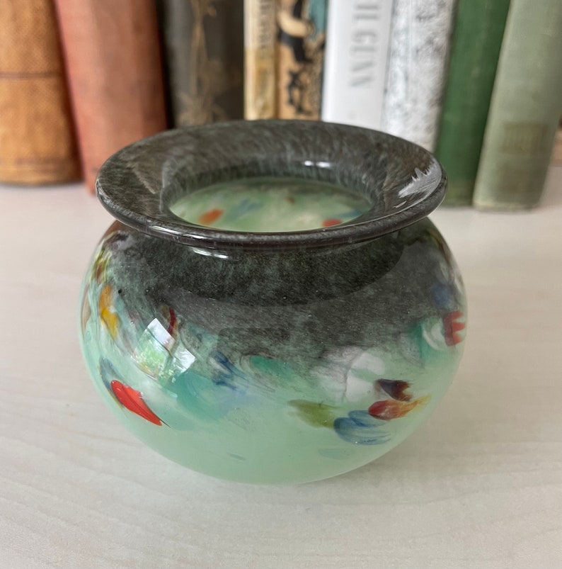 K&ouml;nnte beinhalten: Eine kleine, runde, gr&uuml;ne Glasvase mit einem dunkelgrauen Rand. Die Vase hat einen marmorierten Effekt mit Spritzern in Blau, Rot und Gelb.