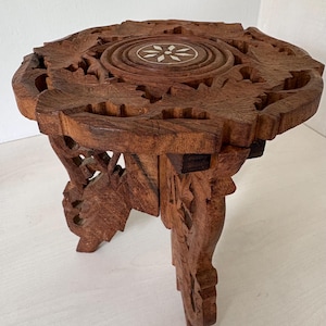 May include: A small, round, brown wooden stool featuring intricate hand-carved floral patterns on the top and legs. The centre of the tabletop displays a circular inlay with a white star-shaped design. The stool has three carved legs.