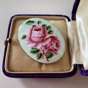 May include: A white oval brooch featuring a hand-painted design of two pink roses with green leaves and stems. The brooch is set in a silver tone metal.