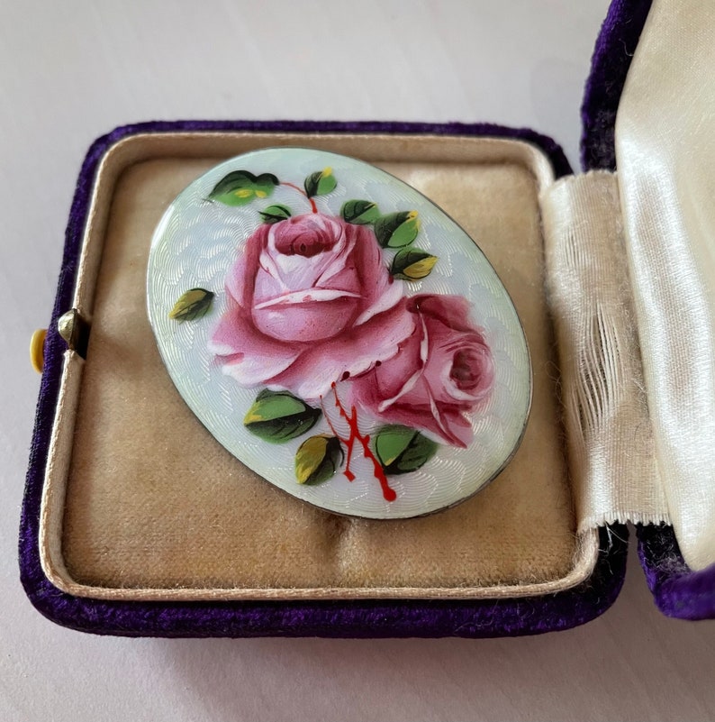 May include: A white oval brooch featuring a hand-painted design of two pink roses with green leaves and stems. The brooch is set in a silver tone metal.