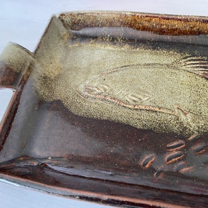 May include: Rectangular ceramic serving tray with a brown and tan glaze. The tray features a raised design of a fish and other aquatic elements. The tray has two handles and is approximately 30 cm long.