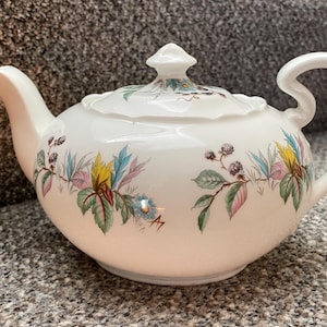 May include: A white ceramic teapot with a curved handle and spout. The teapot is decorated with a floral pattern featuring colourful leaves, berries, and flowers in shades of pink, yellow, and blue. The lid has a small finial.