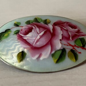 May include: A silver oval brooch with a blue enamel background and hand-painted pink roses with green leaves.