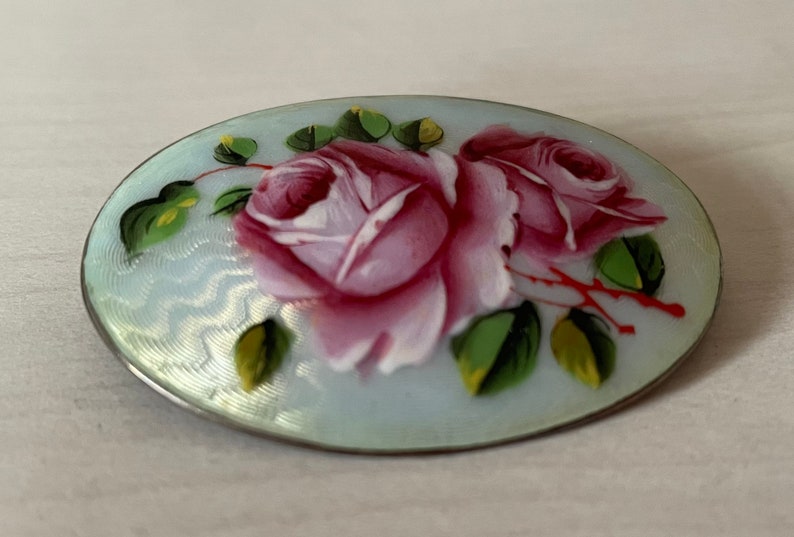 May include: A silver oval brooch with a blue enamel background and hand-painted pink roses with green leaves.