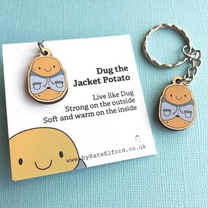 May include: Wooden keychain featuring a cartoon potato wearing a blue jacket. The keychain has a silver key ring and a card with the text "Dug the Jacket Potato. Live like Dug. Strong on the outside. Soft and warm on the inside. www.byKateElford.co.uk"