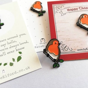 May include: Three enamel pins featuring a robin perched on a branch. The robin is depicted in red, white, and brown with green leaves. The pins are on a white background with a card that says "Happy Christmas".