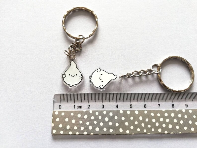 Two silver keychains with clear acrylic charms shaped like flames with black outlines and smiling faces. The charms are approximately 2.5 cm in size. The keychains are on a white surface with a ruler for scale.