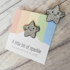 May include: Two glittery star-shaped pins with black outlines and smiling faces. The pins are on a card with a rainbow background and the text "A little bit of sparkle" and the website address.