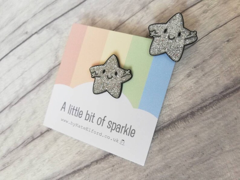 May include: Two glittery star-shaped pins with black outlines and smiling faces. The pins are on a card with a rainbow background and the text "A little bit of sparkle" and the website address.