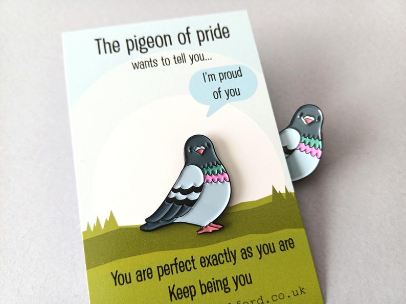 A pin and a card featuring a cartoon pigeon design. The pigeon has a grey head, light blue body, and pink and green accents. The card reads "The pigeon of pride wants to tell you... I'm proud of you."