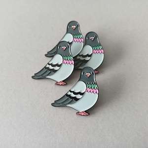 Four pigeon-shaped enamel pins. Each pin has a grey head, light blue body, and black and white wings. The chest features pink and green detailing, and the feet are pink.