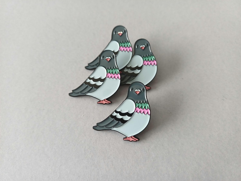 Four pigeon-shaped enamel pins. Each pin has a grey head, light blue body, and black and white wings. The chest features pink and green detailing, and the feet are pink.
