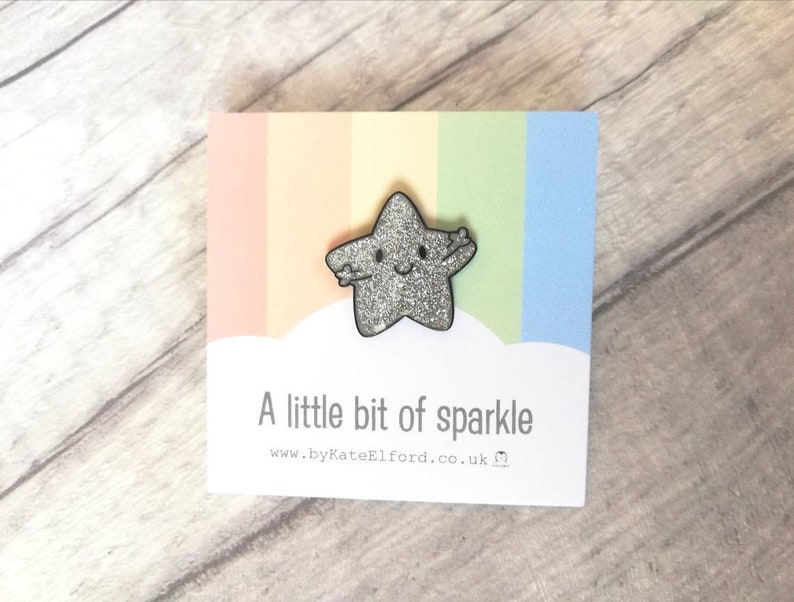 May include: A sparkly silver star-shaped brooch with a smiling face and outstretched arms, presented on a card with a rainbow gradient. The card reads "A little bit of sparkle" and a website address.