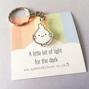 A white flame keyring with a happy face. The keyring is attached to a silver key ring. The keyring is on a card with the text "A little bit of light for the dark".