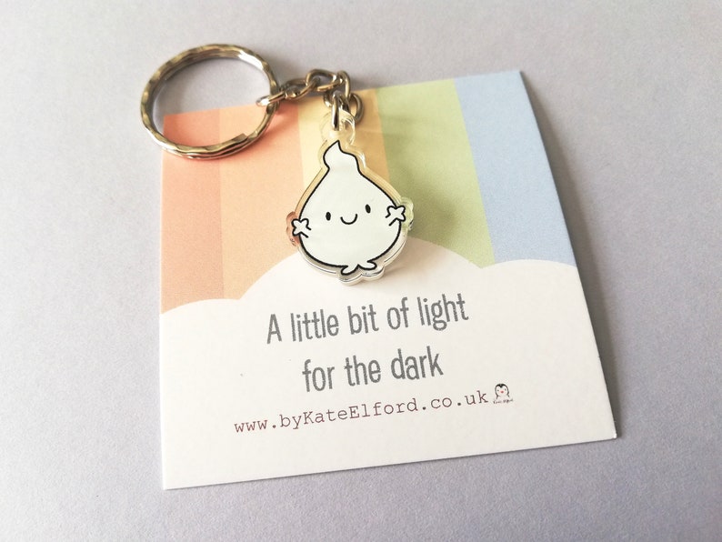 A white flame keyring with a happy face. The keyring is attached to a silver key ring. The keyring is on a card with the text "A little bit of light for the dark".