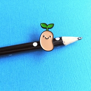 May include: A brown and tan enamel pin of a smiling bean sprout with green leaves. The pin is on a black pencil with white polka dots.