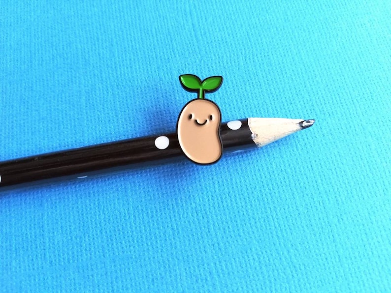 May include: A brown and tan enamel pin of a smiling bean sprout with green leaves. The pin is on a black pencil with white polka dots.