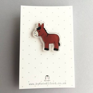 May include: A brown donkey cartoon character on a clear acrylic pin. The pin is on a white card with black polka dots and the text "www.byKateElford.co.uk".
