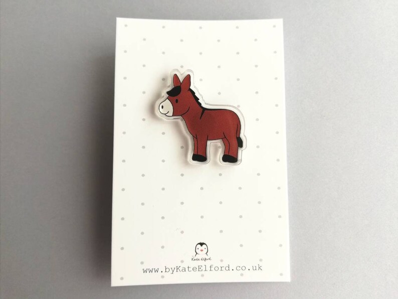 May include: A brown donkey cartoon character on a clear acrylic pin. The pin is on a white card with black polka dots and the text "www.byKateElford.co.uk".