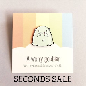 May include: A light green enamel pin of a cartoon character with its tongue sticking out, on a card with the text "A worry gobbler" and "SECONDS SALE". The background has pastel stripes.