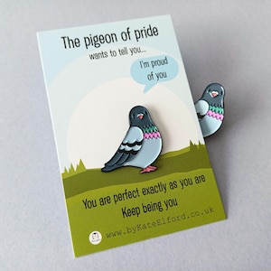A decorative pin in the shape of a pigeon, with blue, grey, and pink details, next to a card. The card says "The pigeon of pride wants to tell you... I'm proud of you. You are perfect exactly as you are. Keep being you."