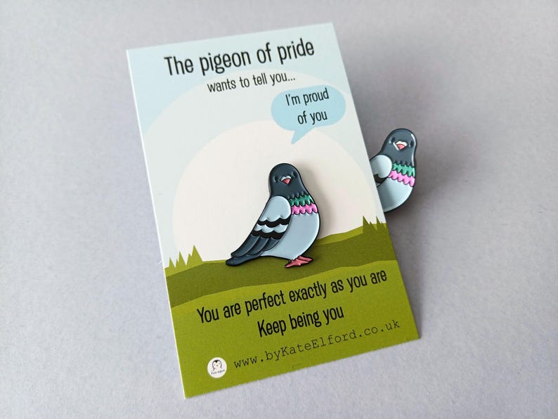 A decorative pin in the shape of a pigeon, with blue, grey, and pink details, next to a card. The card says "The pigeon of pride wants to tell you... I'm proud of you. You are perfect exactly as you are. Keep being you."