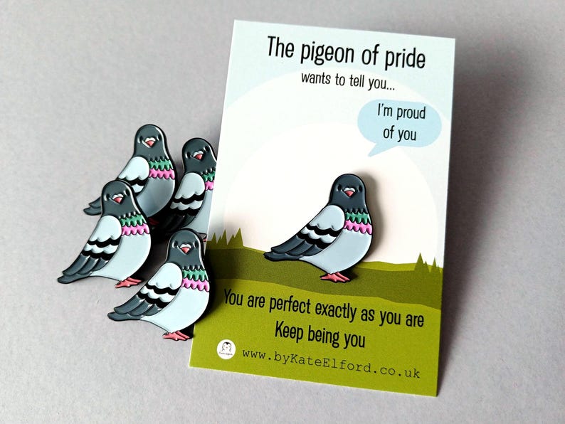 Enamel pins in the shape of pigeons, featuring light blue, grey, pink, and green details. The pins are arranged near a card that reads "The pigeon of pride wants to tell you... I'm proud of you."