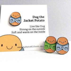May include: Two enamel pins featuring a cartoon potato wearing a jacket. The potato is smiling and has a green or blue jacket with pockets. The text on the card reads "Dug the Jacket Potato", "Live like Dug", "Strong on the outside", "Soft and warm on the inside", and "www.byKateElford.co."