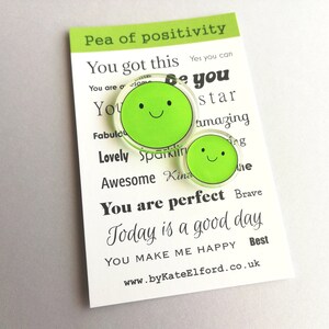 Puede incluir: Dos pines verdes con forma de guisante y caras sonrientes sobre una tarjeta blanca. La tarjeta dice "Pea of positivity" y presenta frases motivadoras como "You got this" y "You are perfect". Tambi&eacute;n es visible la direcci&oacute;n del sitio web www.byKateElford.co.uk.