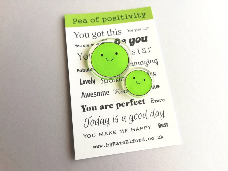 Puede incluir: Dos pines verdes con forma de guisante y caras sonrientes sobre una tarjeta blanca. La tarjeta dice "Pea of positivity" y presenta frases motivadoras como "You got this" y "You are perfect". Tambi&eacute;n es visible la direcci&oacute;n del sitio web www.byKateElford.co.uk.