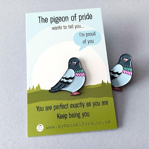 Two enamel pins depicting a cartoon pigeon with blue, black, pink, and red details. The pins are placed near a card that reads "The pigeon of pride wants to tell you... I'm proud of you. You are perfect exactly as you are. Keep being you."