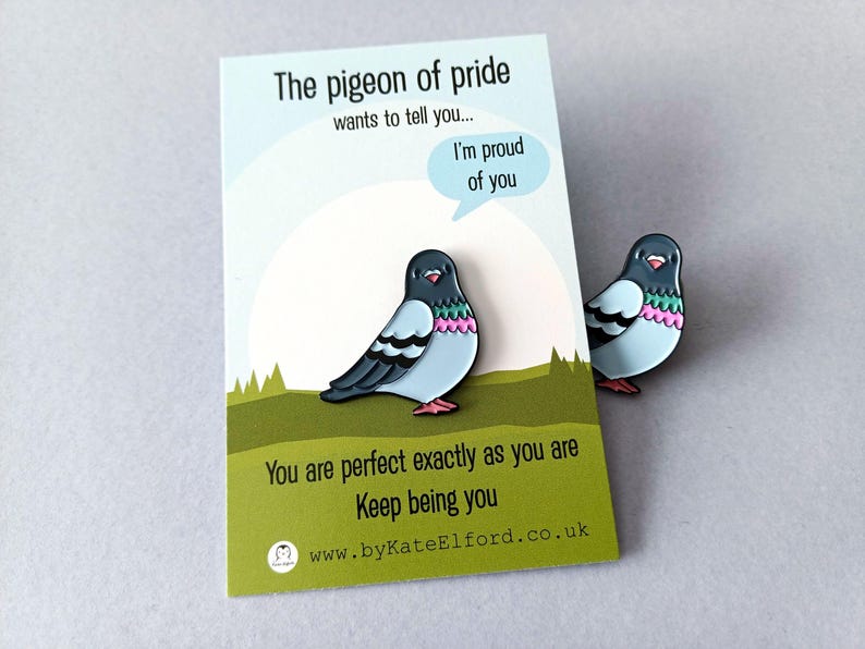 Two enamel pins depicting a cartoon pigeon with blue, black, pink, and red details. The pins are placed near a card that reads "The pigeon of pride wants to tell you... I'm proud of you. You are perfect exactly as you are. Keep being you."