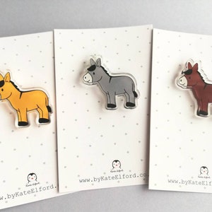 May include: Three donkey pins, each with a different color: yellow, gray, and brown. The pins are made of clear acrylic and have a white background. The website address "www.byKateElford.co.uk" is printed on the bottom of each card.