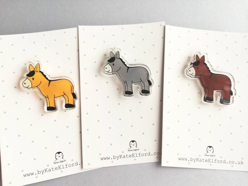 May include: Three donkey pins, each with a different color: yellow, gray, and brown. The pins are made of clear acrylic and have a white background. The website address "www.byKateElford.co.uk" is printed on the bottom of each card.