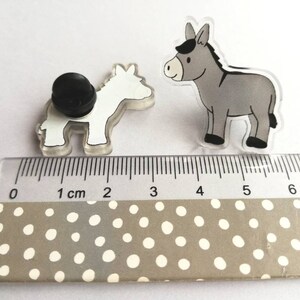 May include: Two donkey pins, one gray and one white, are shown on a ruler with a white background. The gray donkey is 2.5 inches tall and the white donkey is 2 inches tall.