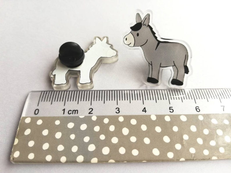 May include: Two donkey pins, one gray and one white, are shown on a ruler with a white background. The gray donkey is 2.5 inches tall and the white donkey is 2 inches tall.