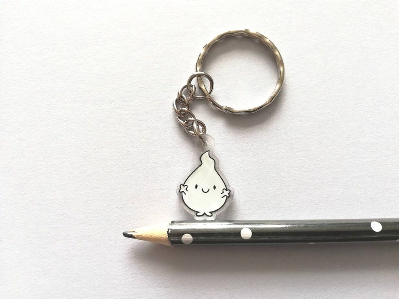 A white flame keyring with a smiling face and a silver key ring. The keyring is hanging from a pencil with black and white polka dots.