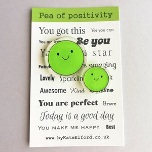 Puede incluir: Dos pines verdes con forma de guisante y caras sonrientes se muestran en una tarjeta blanca. La tarjeta tiene el texto "Pea of positivity" y frases inspiradoras como "You got this" y "Today is a good day". Tambi&eacute;n se ve la direcci&oacute;n del sitio web www.byKateElford.co.uk.
