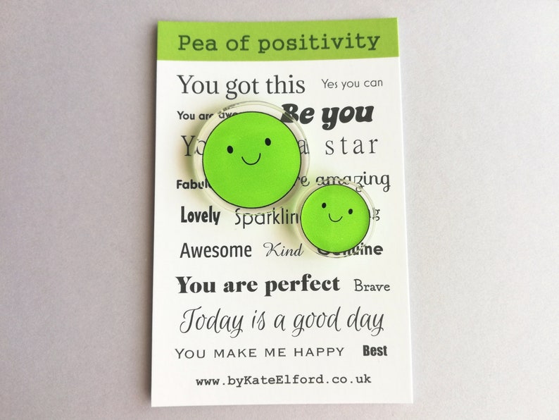 Puede incluir: Dos pines verdes con forma de guisante y caras sonrientes se muestran en una tarjeta blanca. La tarjeta tiene el texto "Pea of positivity" y frases inspiradoras como "You got this" y "Today is a good day". Tambi&eacute;n se ve la direcci&oacute;n del sitio web www.byKateElford.co.uk.