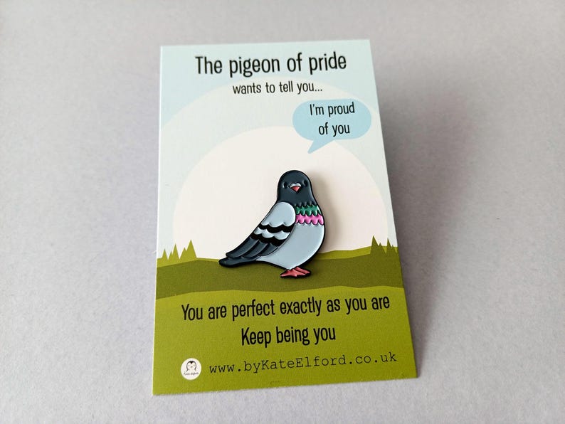 A colourful pigeon enamel pin on a backing card. The card reads "The pigeon of pride wants to tell you... I'm proud of you." and "You are perfect exactly as you are. Keep being you." The pin is a cute, positive design.